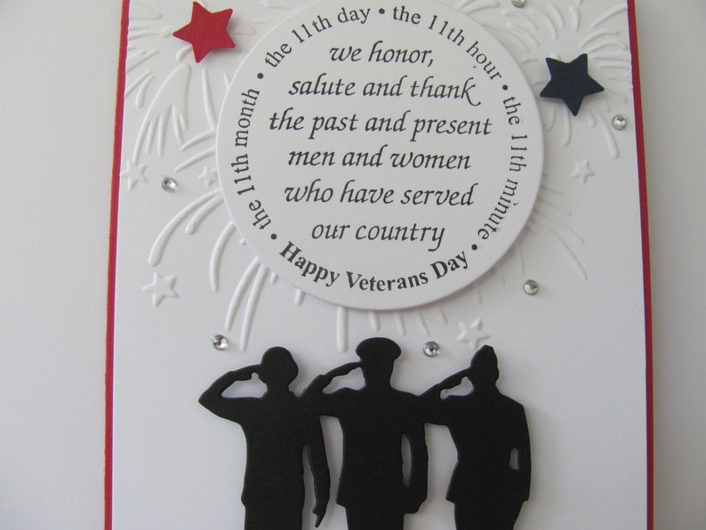 Pu&ograve; includere: Un biglietto bianco per il Veterans Day con un bordo rosso. La carta presenta un design circolare con testo in onore dei veterani, circondato da stelle e fuochi d'artificio. Sagome di soldati che salutano in basso.