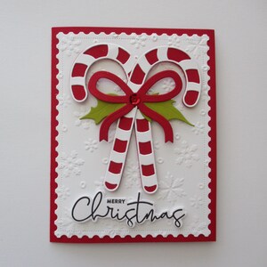 K&ouml;nnte beinhalten: Eine Weihnachtskarte mit zwei rot-wei&szlig; gestreiften Zuckerstangen, einer roten Schleife und gr&uuml;nen Stechpalmenbl&auml;ttern. Der wei&szlig;e Hintergrund hat gepr&auml;gte Schneeflocken und den Schriftzug "Merry Christmas".