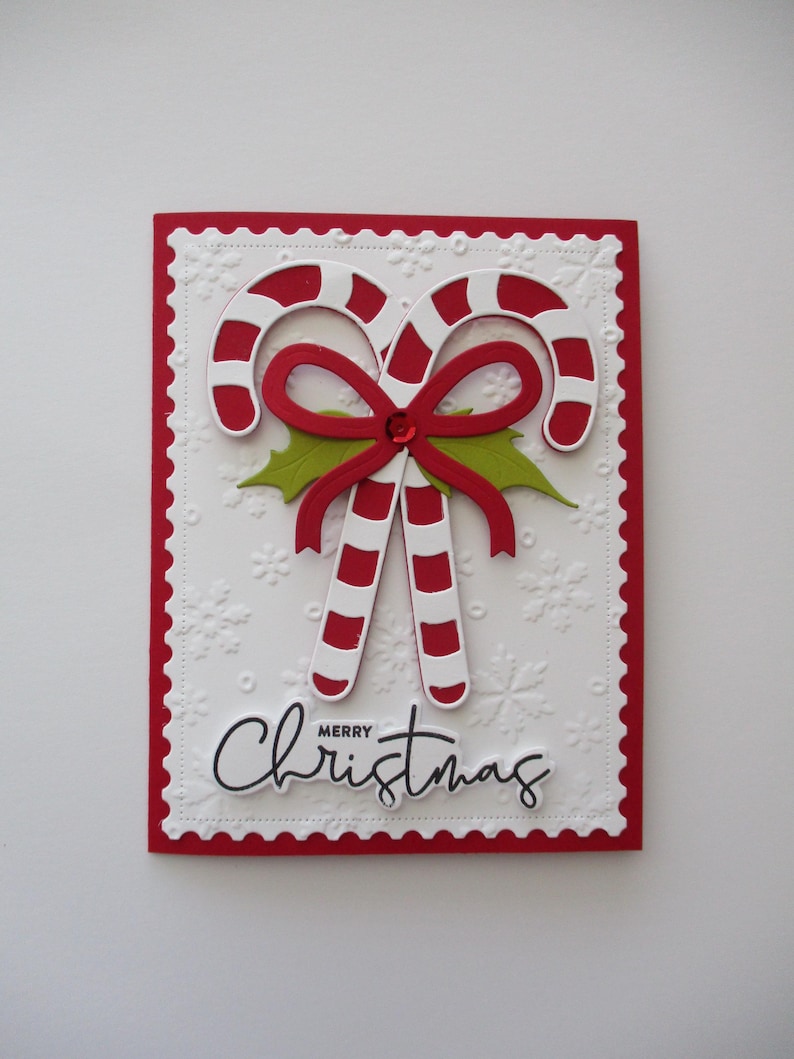 K&ouml;nnte beinhalten: Eine Weihnachtskarte mit zwei rot-wei&szlig; gestreiften Zuckerstangen, einer roten Schleife und gr&uuml;nen Stechpalmenbl&auml;ttern. Der wei&szlig;e Hintergrund hat gepr&auml;gte Schneeflocken und den Schriftzug "Merry Christmas".