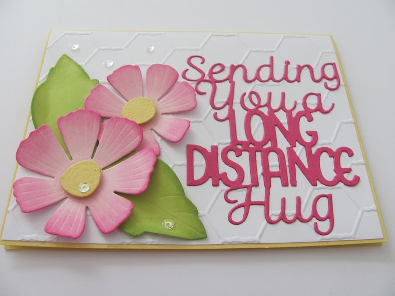May include: A white card with a pink and green floral design. The card has the text "Sending you a long distance hug" in pink.