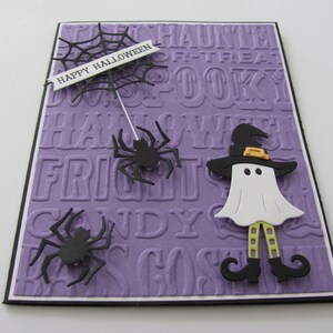 Może przedstawiać: Fioletowa kartka z czarną pajęczyną, duchem w czarodziejskim kapeluszu i dwoma czarnymi pająkami. Na kartce widnieje napis "Happy Halloween" na białym banerze.