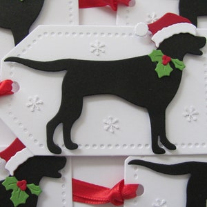 Pu&ograve; includere: Etichette regalo bianche con una silhouette nera di un cane che indossa un cappello di Babbo Natale e agrifoglio. Le etichette hanno un bordo punteggiato e disegni di fiocchi di neve.