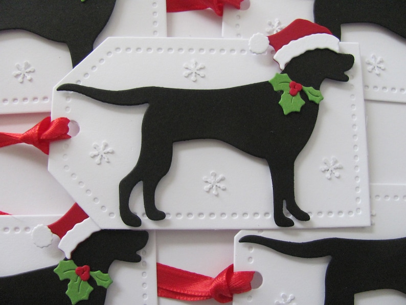 Pu&ograve; includere: Etichette regalo bianche con una silhouette nera di un cane che indossa un cappello di Babbo Natale e agrifoglio. Le etichette hanno un bordo punteggiato e disegni di fiocchi di neve.