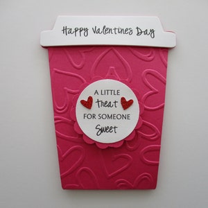 May include: A Valentine's Day card shaped like a coffee cup, in shades of pink and white. The cup has a heart-shaped pattern and the words "Happy Valentine's Day" at the top. A white circle in the middle says "A little treat for someone sweet."