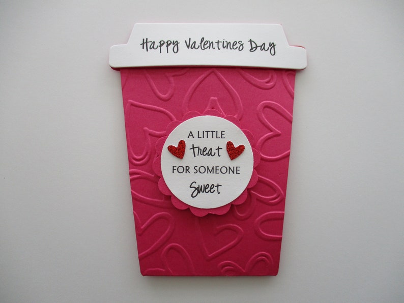 May include: A Valentine's Day card shaped like a coffee cup, in shades of pink and white. The cup has a heart-shaped pattern and the words "Happy Valentine's Day" at the top. A white circle in the middle says "A little treat for someone sweet."