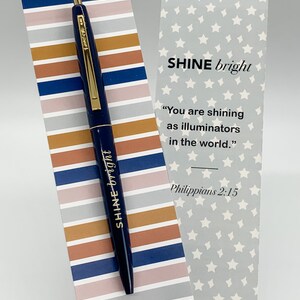 May include: A blue pen with gold accents and the words "SHINE bright" printed on it. The pen is on a striped background with the words "SHINE bright" printed on it. The background is a mix of brown, pink, and blue stripes. The pen is next to a bookmark with a grey background and white stars. The bookmark has the words "SHINE bright" printed on it, along with a quote from Philippians 2:15. The bookmark also has the words "connect et.net/giftshop" printed on it.