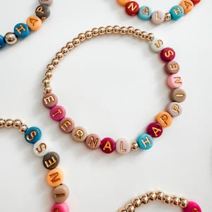 May include: A beaded bracelet featuring metallic gold-tone spherical beads interspersed with matte-finish round beads in shades of pink, blue, orange, and beige. The beads are imprinted with gold letters spelling out the phrase ETERNAL HAPPINESS.
