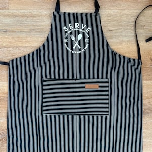 Peut inclure: Tablier à rayures noires et blanches avec une grande poche avant et une étiquette en cuir. Le tablier porte l'inscription "SERVE ONE ANOTHER WITH LOVE" dans un motif circulaire avec des ustensiles croisés.