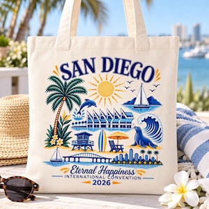 May include: A cream-colored canvas tote bag featuring a colorful graphic print with the text SAN DIEGO, Eternal Happiness, INTERNATIONAL CONVENTION, and 2026. The design includes a palm tree, sun, sailboat, dolphin, sea lion, and bridge.