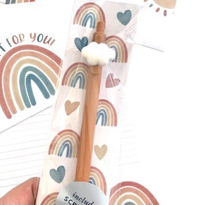 May include: A pen with a cloud-shaped topper is packaged with a bookmark. The pen is light brown, and the bookmark features rainbow and heart designs in muted colors. The packaging includes the text "includes SCRIPTURE bookmark". The background includes the text "GIFT FOR YOU!"