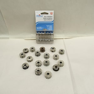 May include: A collection of sewing machine bobbins, both loose and in a package. The package, labeled "PRYM", contains 12 bobbins. The bobbins are silver-colored metal with a central hole and a series of smaller holes around the perimeter. One bobbin is a darker brown color.
