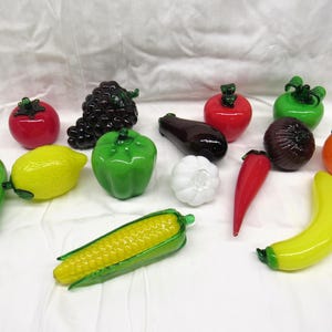May include: A collection of glass fruit and vegetables, including a green pepper, a red tomato, a yellow banana, a green zucchini, a purple eggplant, a red apple, a green grape bunch, a yellow corn cob, a white onion, a yellow lemon, and an orange.