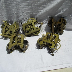 May include: A collection of five vintage brass clock movements featuring intricate gear systems, metal plates, and coiled springs. These mechanical components are exposed, showing the internal structure of timekeeping mechanisms with various cogs.