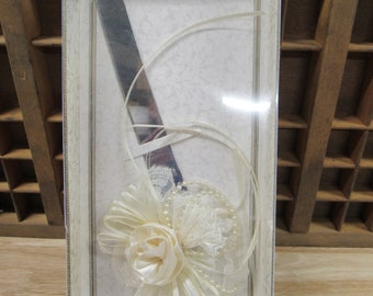 May include: A white and silver cake knife with a white satin bow and pearl accents. The knife is in a clear plastic box with a white background and the text "Lillian Rose" in gold lettering. The text below the knife reads "In the language of the pure and true, a rose is an expression of love. Lillian Rose - a pure and true expression of love."