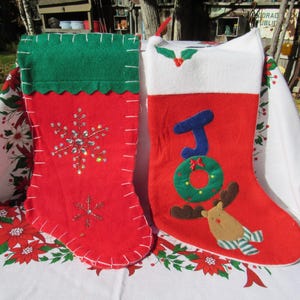 May include: Two Christmas stockings. One red stocking has a green cuff and a snowflake design with sequins. The other red stocking has a white cuff, the word "JOY", a wreath, and a reindeer design. Both stockings are on a white tablecloth.