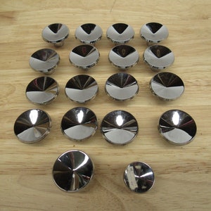 May include: A collection of shiny, silver-colored cabinet knobs arranged on a light brown wooden surface. The knobs have a circular, domed design with a reflective finish, creating a modern aesthetic. The knobs are likely for furniture or cabinetry.