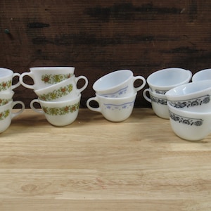 May include: A set of vintage white ceramic mugs with floral patterns. The mugs have a blue and white floral pattern, a green and brown floral pattern, and a black and white floral pattern.