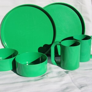 May include: A set of bright green plastic tableware, including two large plates, two mugs with square handles, and two small bowls. The items are arranged on a white surface, showcasing a retro picnic or camping theme.
