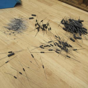 May include: An assortment of electronic components, including resistors and diodes, are scattered on a light wood surface. The components have black cylindrical bodies with wire leads, and some are grouped together.