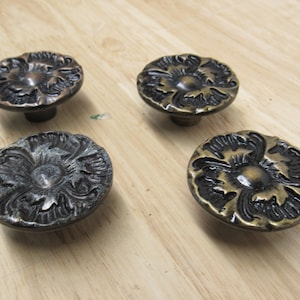 May include: Four antique bronze cabinet knobs with a floral design. The knobs have a detailed, raised pattern and a dark, aged finish. They are arranged on a light wood surface.