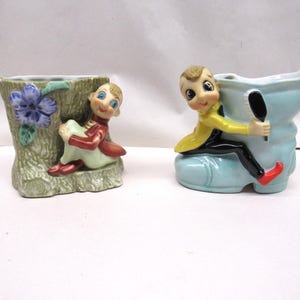 May include: Two ceramic planter figurines of elves. One elf is sitting on a tree stump with a flower. The other elf is sitting in a blue boot.