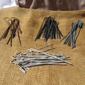 May include: A collection of U-shaped metal garden stakes arranged in four distinct groups. One group is rusty brown, one is matte black, one is dark grey, and a larger pile in the foreground consists of light grey, galvanized metal stakes.