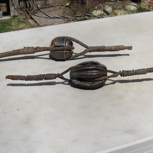 May include: Two antique metal pulleys with thick, twisted wire rope. One pulley has a rectangular cage, the other a ribbed, rounded shape. Both are weathered with a rusty brown color, suggesting age and outdoor use. The pulleys are set against a light background.
