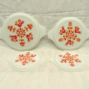 Puede incluir: Un juego de cuatro tapas blancas para platos Pyrex. Cada tapa presenta un diseño floral central con detalles en rojo y amarillo, y motivos de pájaros. Las tapas son redondas con asas, ideales para cocina vintage.