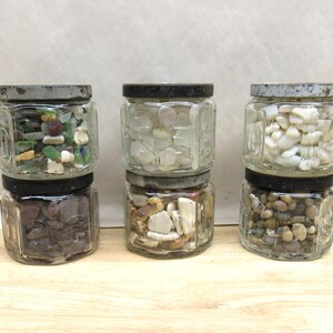May include: Six glass jars filled with various decorative stones and sea glass. The jars have black lids and are stacked in pairs. The stones include shades of brown, white, green, and clear.