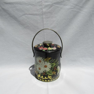 May include: A cylindrical black metal canister featuring a painted floral design with white daisies, green leaves, and red accents. The container has a removable lid with a hexagonal knob and a thin, arched metal handle attached to the sides.