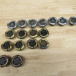 May include: A collection of decorative buttons in various metallic finishes, including silver, bronze, and gold. Each button features a spiral design in the center and a textured outer edge. The buttons are arranged on a light wood surface.