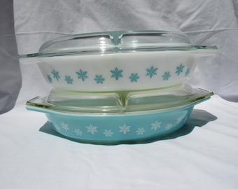 May include: Two stacked oval glass casserole dishes with clear glass lids. The top dish is white with a repeating pattern of light blue snowflakes. The bottom dish is light blue with a repeating pattern of white snowflakes around the exterior.