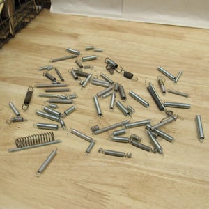 May include: A collection of various metal springs scattered on a light wood surface. The springs vary in size and shape, including coil, extension, and compression springs. Some have loops or hooks at the ends.
