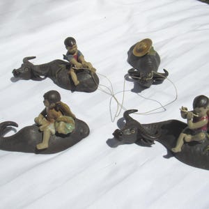 May include: Four ceramic figurines of people riding water buffalo. The figurines are brown and black with painted details. The people are wearing colorful clothing and hats.