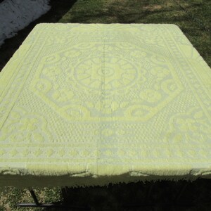May include: A light yellow rectangular tablecloth with a detailed floral pattern. The tablecloth is made of a textured fabric and has a fringed edge. The tablecloth is placed on a table outdoors.