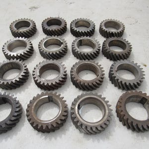 May include: A collection of sixteen metal gears with a circular design and toothed edges. The gears vary in color from silver to brown, suggesting different levels of wear or material composition. The gears are arranged on a light-colored surface.