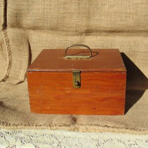 May include: A rectangular wooden box with a brass handle and clasp. The box is stained a warm reddish-brown color. The handle is arched, and the clasp is rectangular. The box is sitting on a burlap surface.