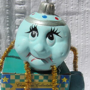 May include: A whimsical Christmas ornament featuring a light blue face with black eyelashes and a smiling red mouth. The ornament has a silver cap and is attached to a box with the text "Christmas Collectibles". Gold tinsel accents the design.