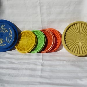 May include: A collection of colorful Tupperware lids in various sizes and shades. The lids include blue, yellow, green, and orange. One blue lid features the text "Let Freedom Ring" and the year "1976".