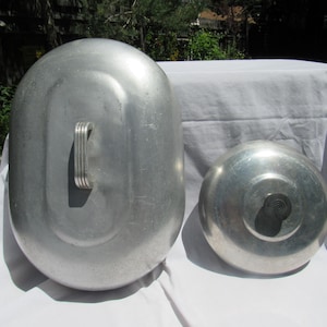 May include: An oval-shaped aluminum lid with a ribbed metal handle and a smaller, circular aluminum lid featuring a black, tiered knob. Both lids have a smooth, metallic finish and are positioned side-by-side on a white fabric surface.