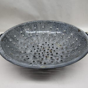 May include: A gray and white enamel colander with a speckled pattern and holes throughout. The colander has two handles on the sides.
