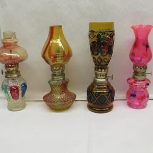 May include: Four antique oil lamps with glass shades and metal bases. The lamps feature various colors, including red, yellow, pink, and green. The lamps have decorative patterns and are of different shapes and sizes.