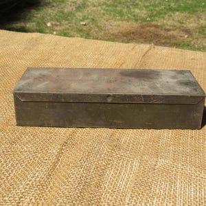 May include: A rectangular metal box with a hinged lid. The box has a weathered, aged appearance with a muted green-gray color. The box is sitting on a tan burlap surface.
