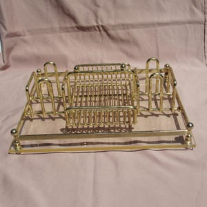 May include: A rectangular gold-tone metal vanity tray featuring a clear glass base. The tray includes a central wire basket compartment flanked by two upright arched wire holders on each side, all finished with decorative ball-topped corner posts.