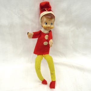 May include: A red and white felt elf doll with yellow legs and a painted face. The elf is wearing a red hat with a white trim.