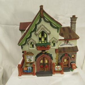 May include: A decorative ceramic miniature building with a sign that reads "Needlework". The building has a brown roof, green trim, and a sign that reads "Hand Made Quilts". The building is decorated with wreaths and snow.