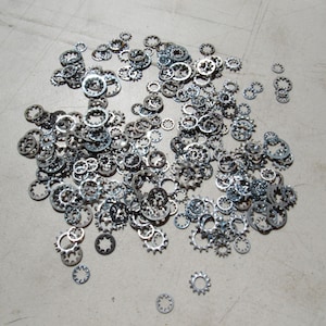 May include: A collection of various sizes of silver-colored, toothed lock washers scattered on a light gray surface. The washers have a circular shape with a series of teeth around the outer edge and inner hole.