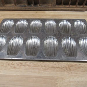May include: A silver metal baking pan with 12 individual molds shaped like seashells. The pan is designed for making madeleines.