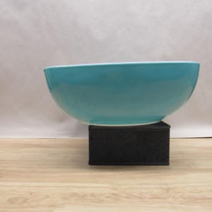 May include: A turquoise blue ceramic bowl with a smooth, rounded shape. The bowl is sitting on a black rectangular base.