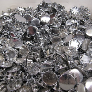 May include: A large pile of silver metal snap fasteners. The fasteners are round and have a raised center with a small hole in the middle. They are used to attach fabric together.